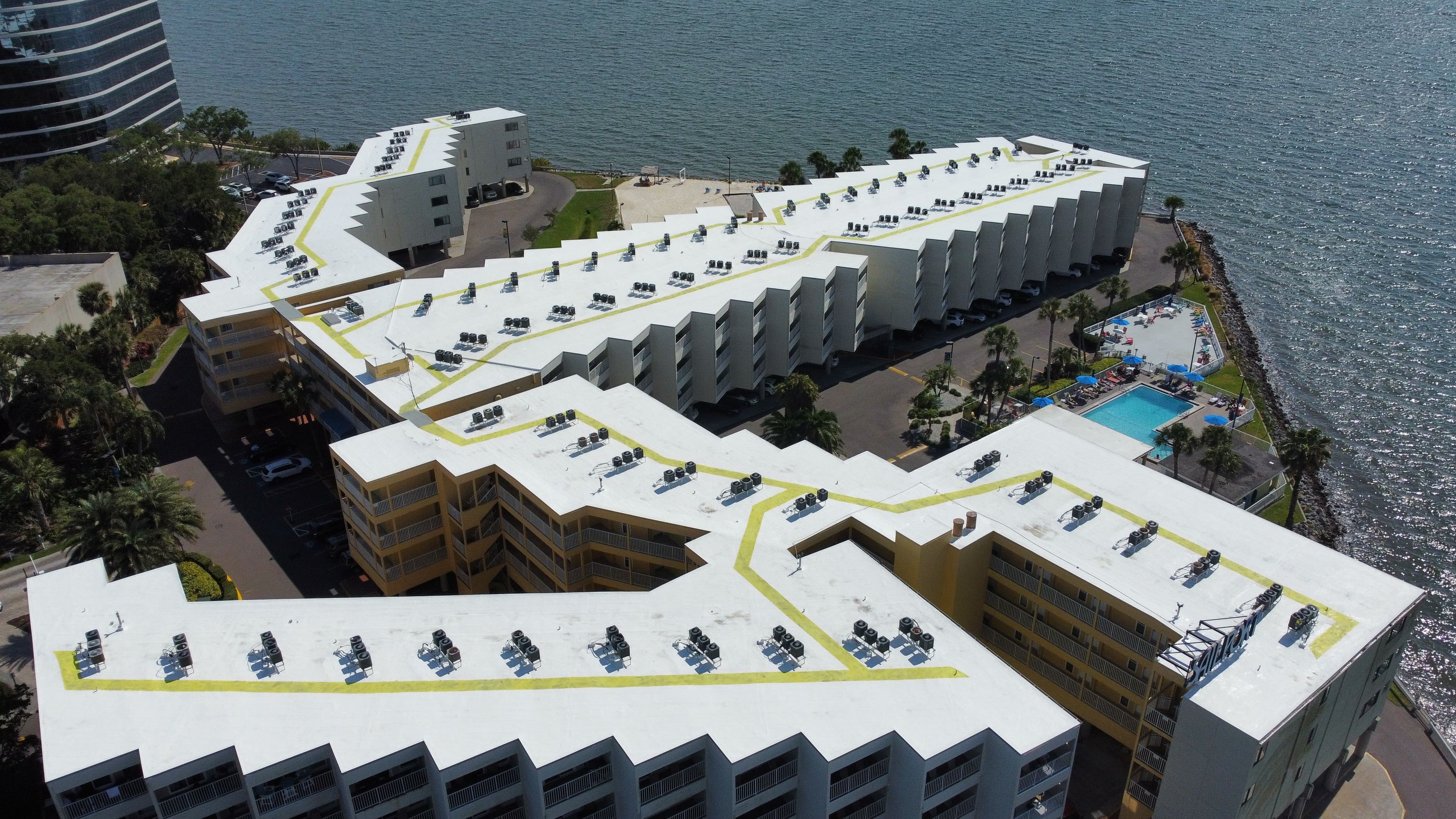 Aerial view of a waterfront hotel complex with a pool and sun loungers near the water.