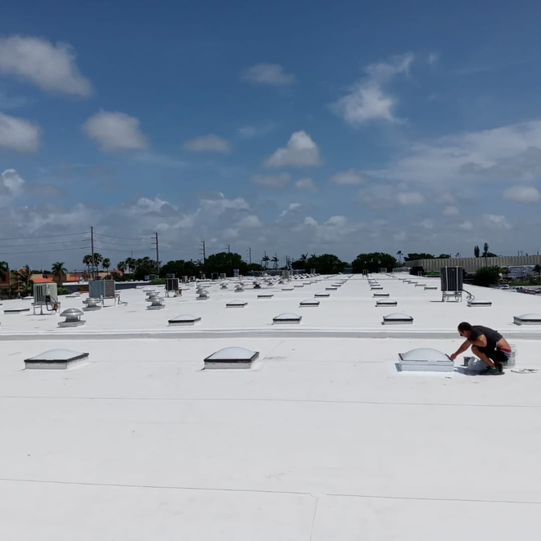 Person inspecting a white rooftop with skylights under a blue sky.