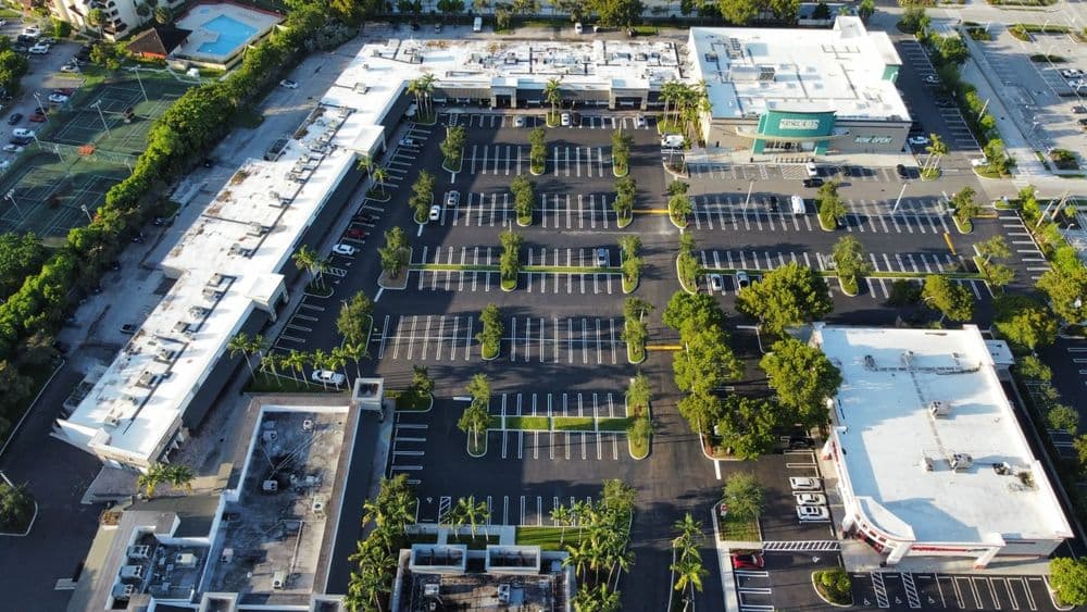 Aerial view of a large shopping center parking lot with palm trees and nearby tennis courts.