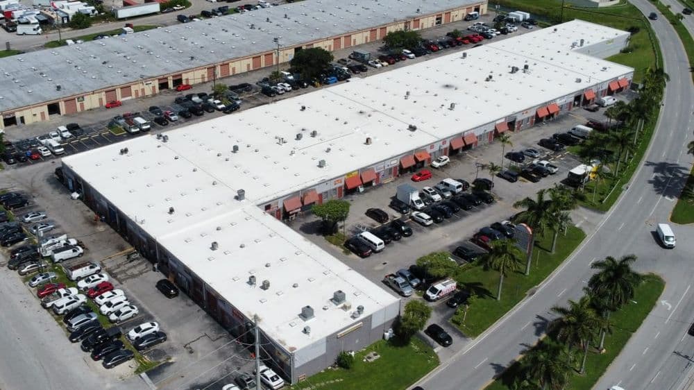 Aerial view of a commercial building with parking lot and palm trees nearby.
