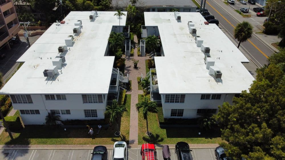Aerial view of two apartment buildings with white roofs and lush greenery between them.
