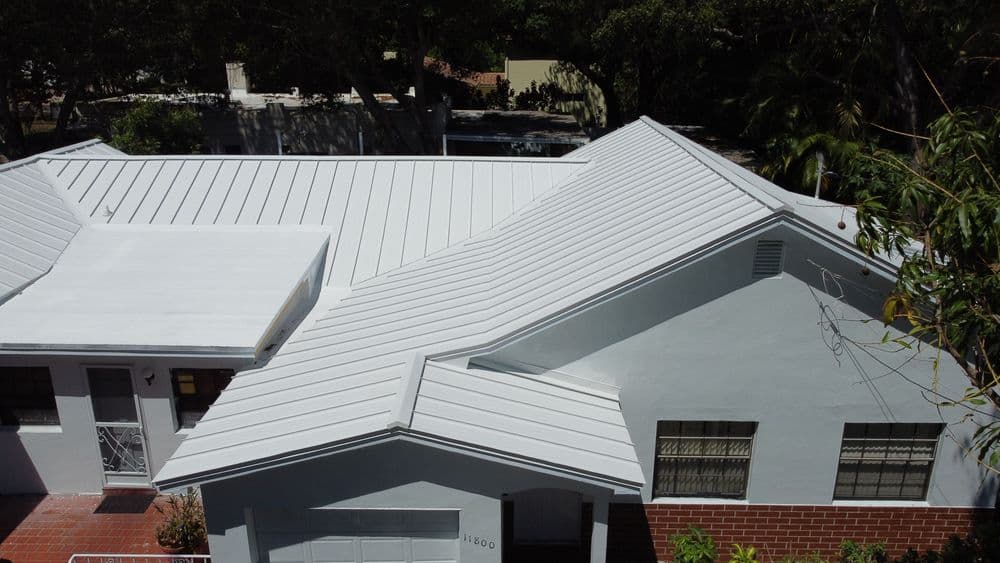 Modern residential house with a white metal roof and light-colored exterior, surrounded by trees.