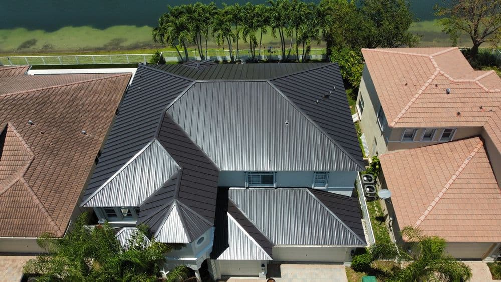 Aerial view of a modern home with a sleek metal roof and lush landscaping by a water body.