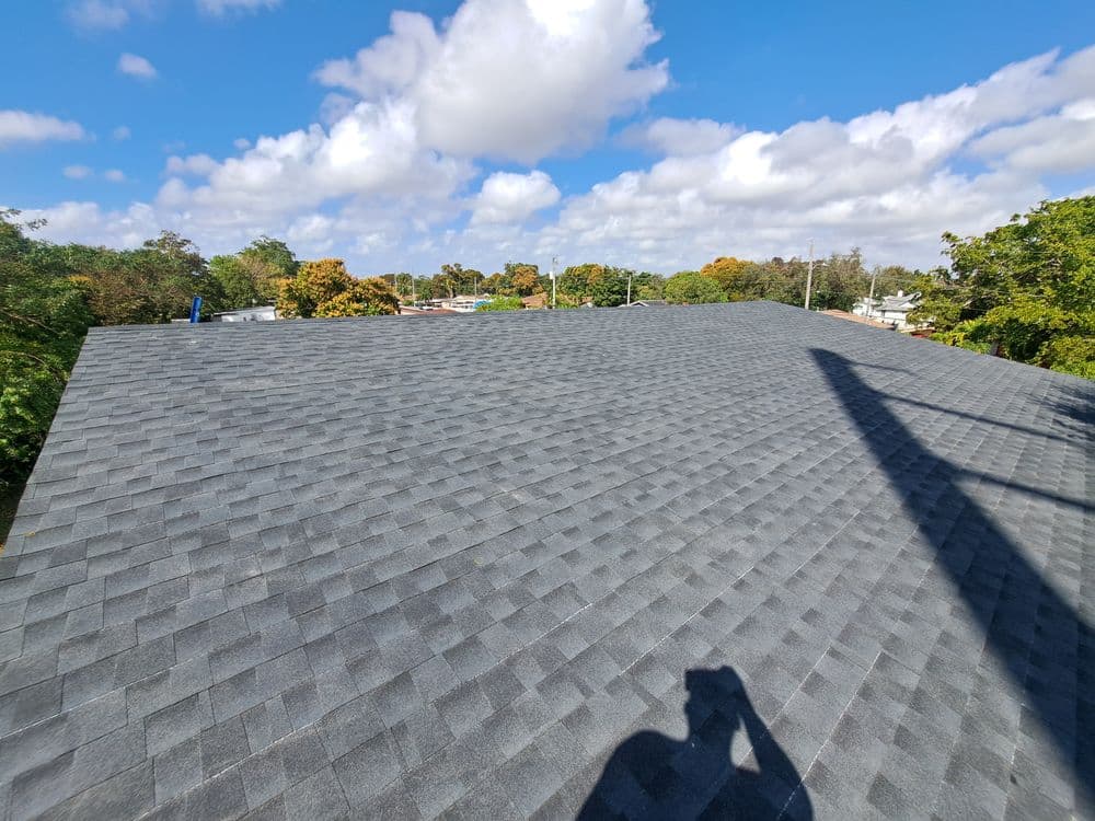 Flat gray asphalt shingle roof under a blue sky with scattered clouds and tree foliage.