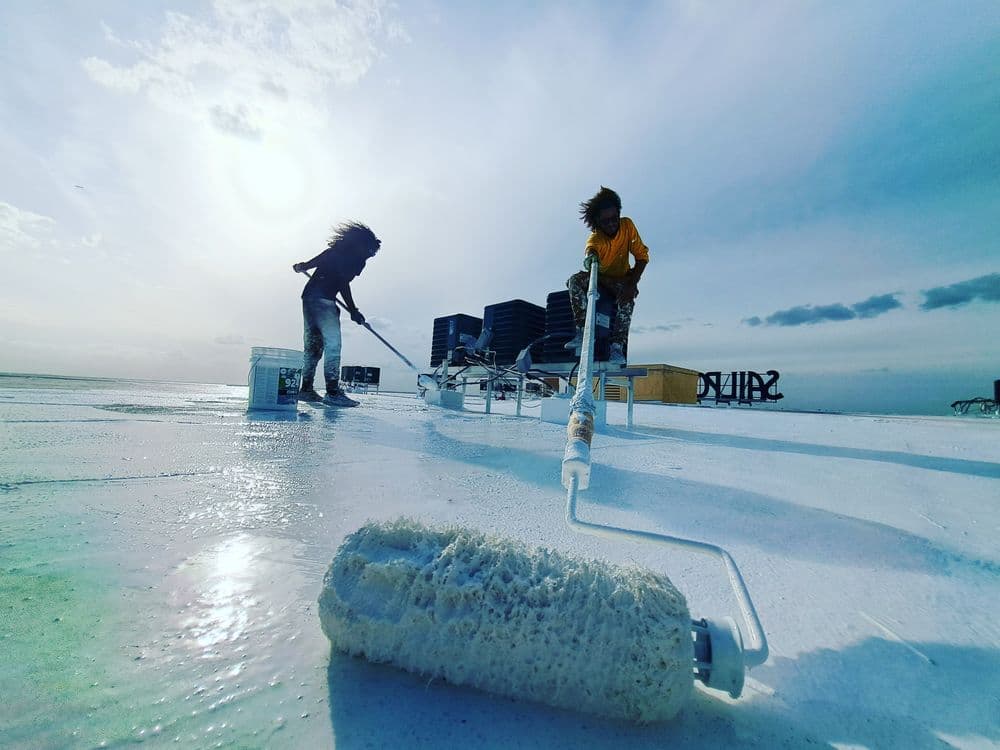 Workers applying white roof coating with paint rollers on a sunny day.