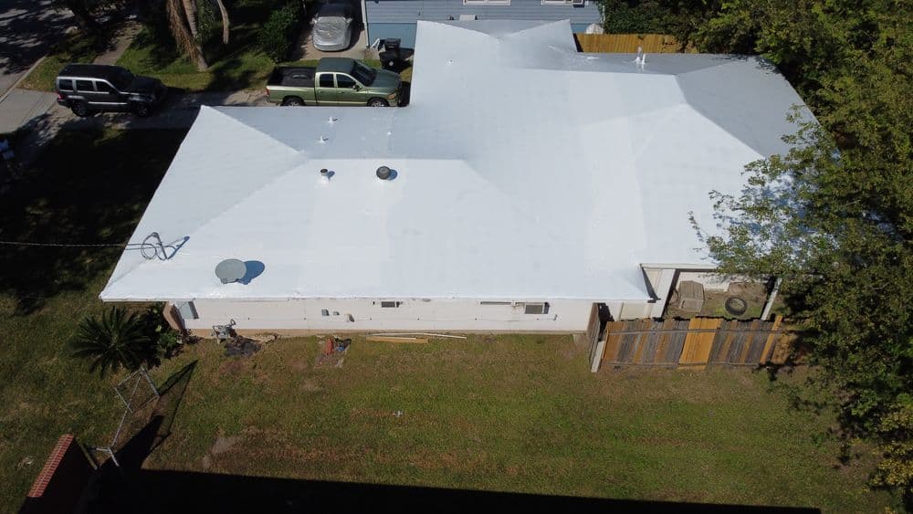 Aerial view of a residential home with a white roof and backyard, surrounded by greenery.